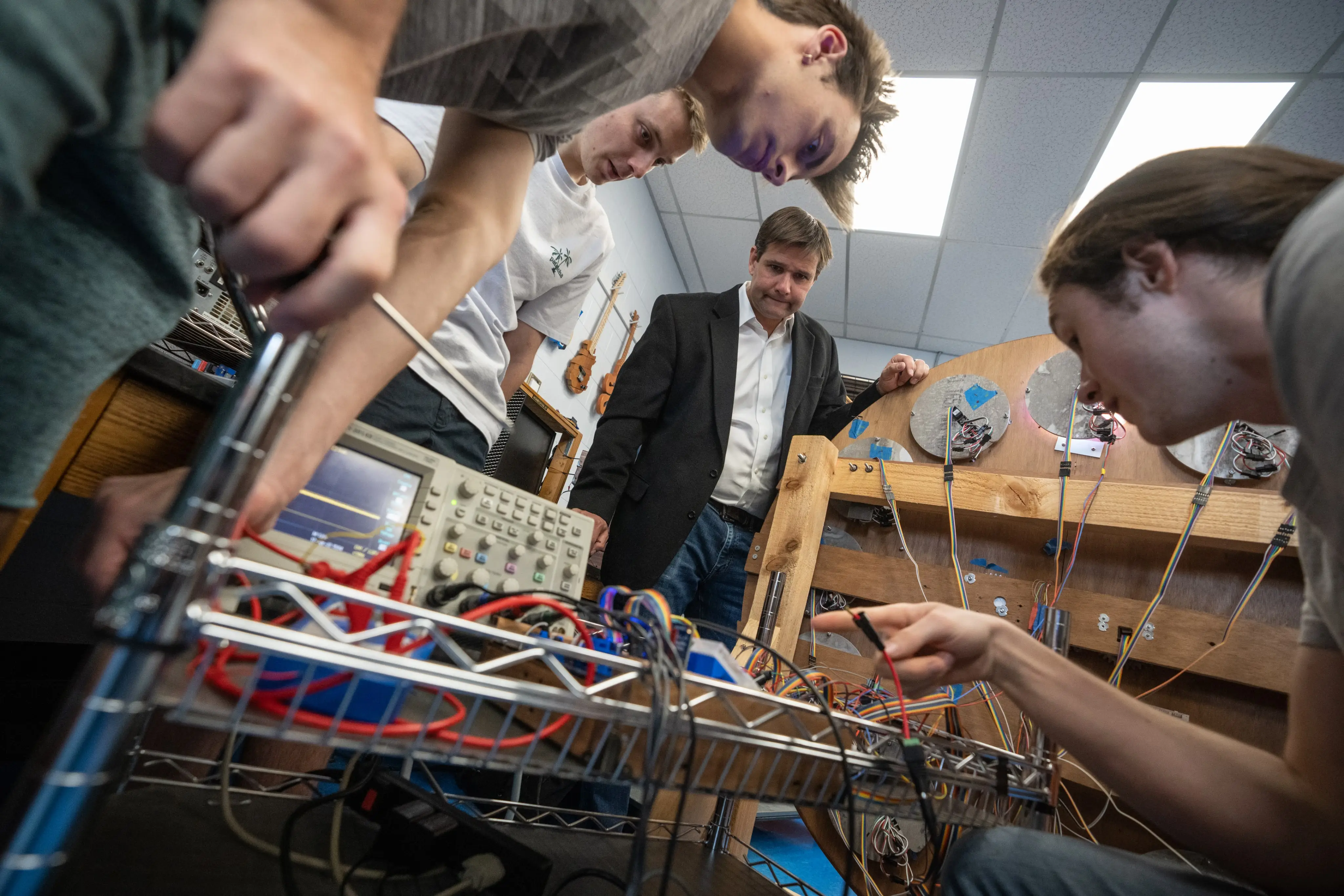 A group examines electronic equipment and wiring on a rack in a lab setting