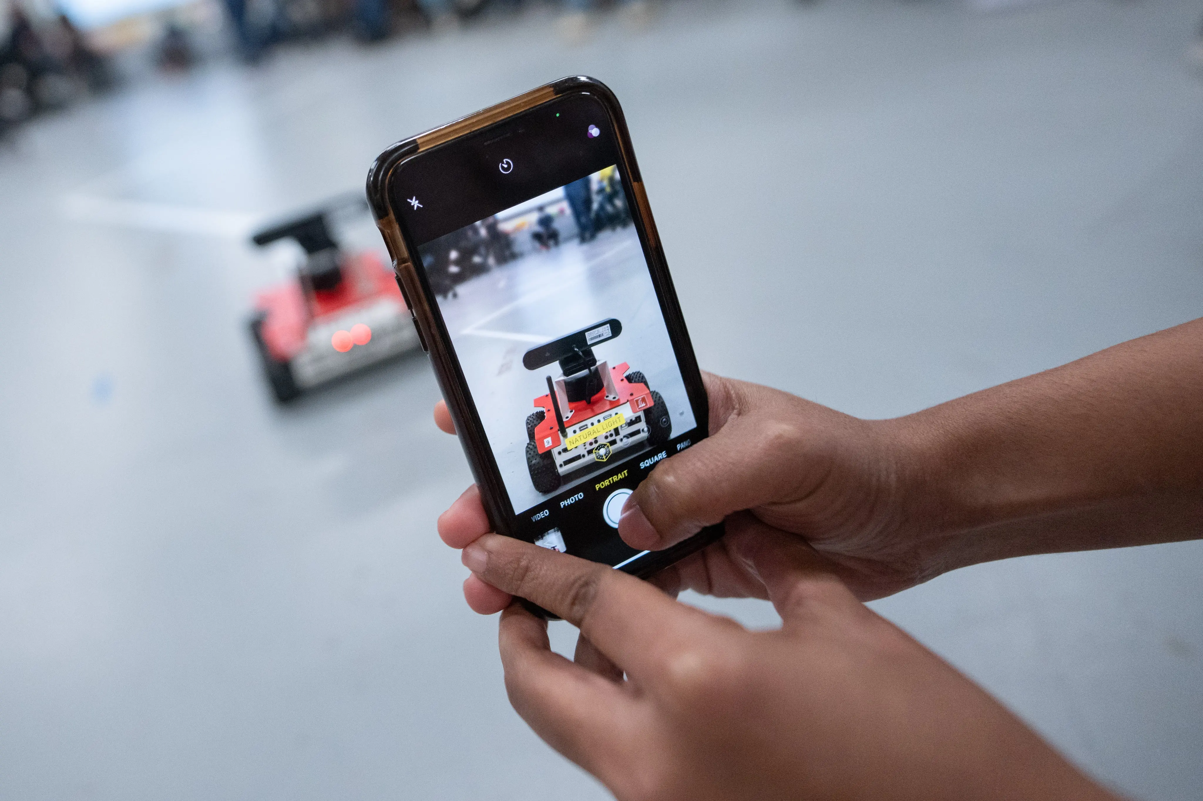 A person taking a photo with a smartphone of a small red robotic device with wheels.