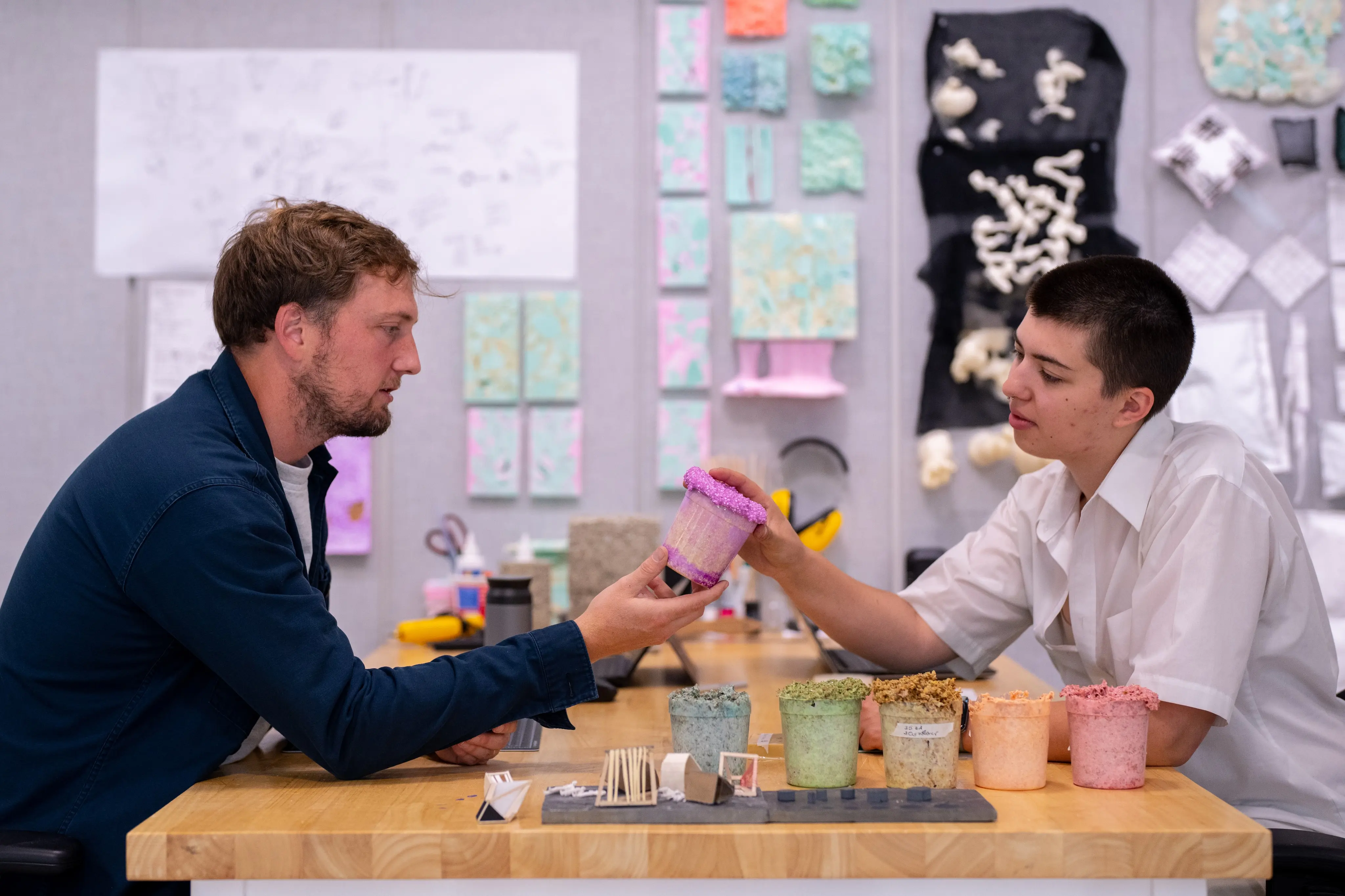 Two people sitting at a table in an art studio examine colorful textured containers