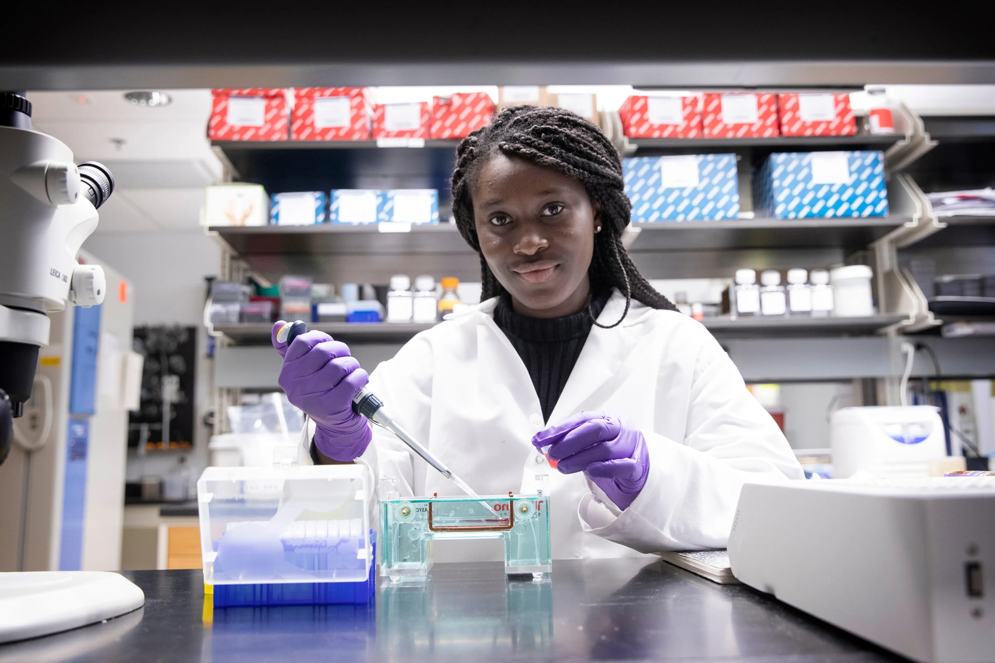 A scientist in a lab coat and purple gloves uses a pipette while working with laboratory equipment